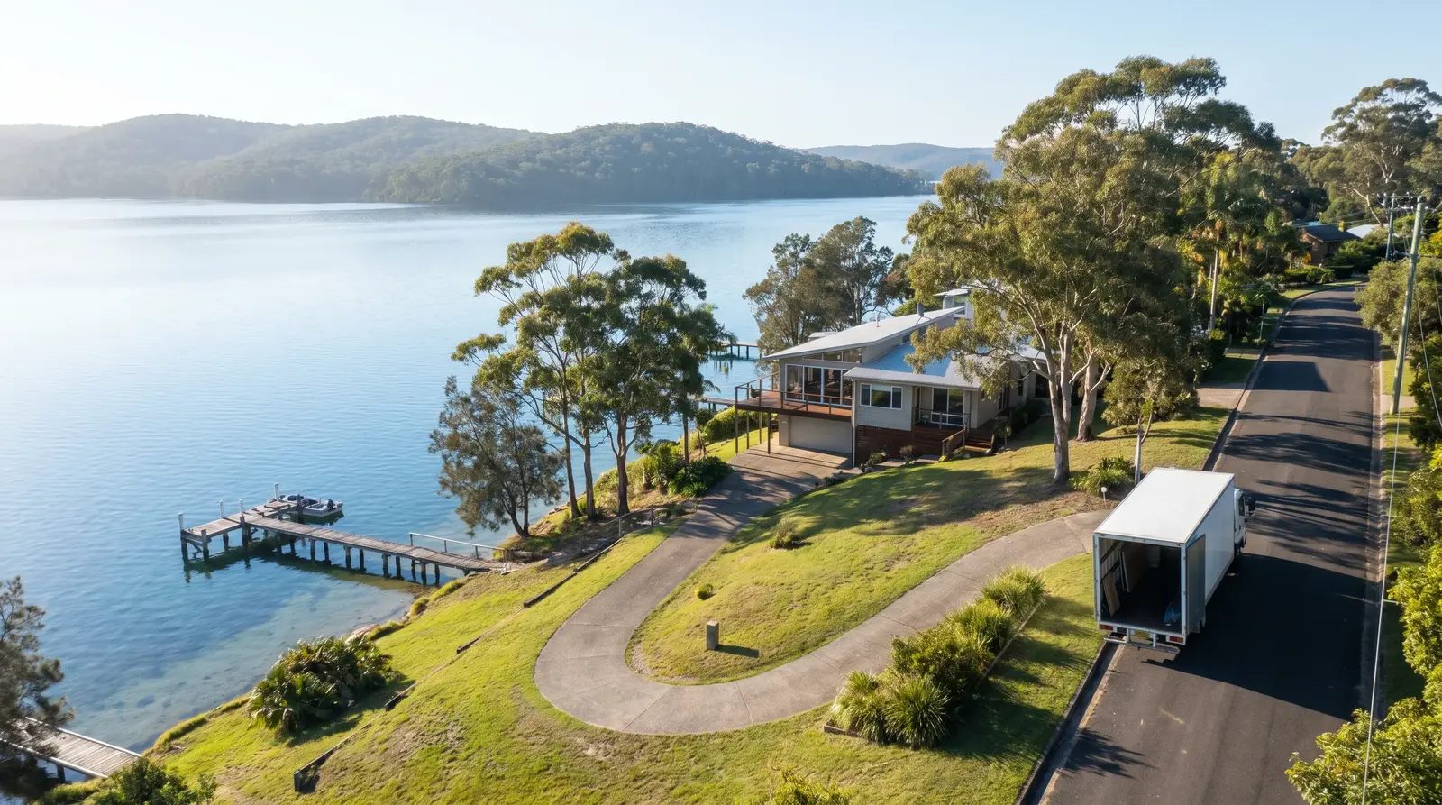 A Lake Macquarie waterfront home with a long sloping driveway down to a private jetty on the calm lake, removal truck parked at the top of the drive